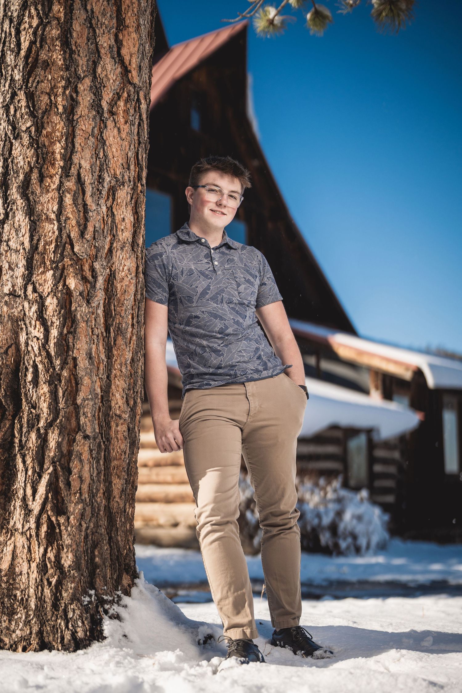Teen boy in glasses leaning against pine tree in snow, wearing khaki pants and gray polo near log cabin.