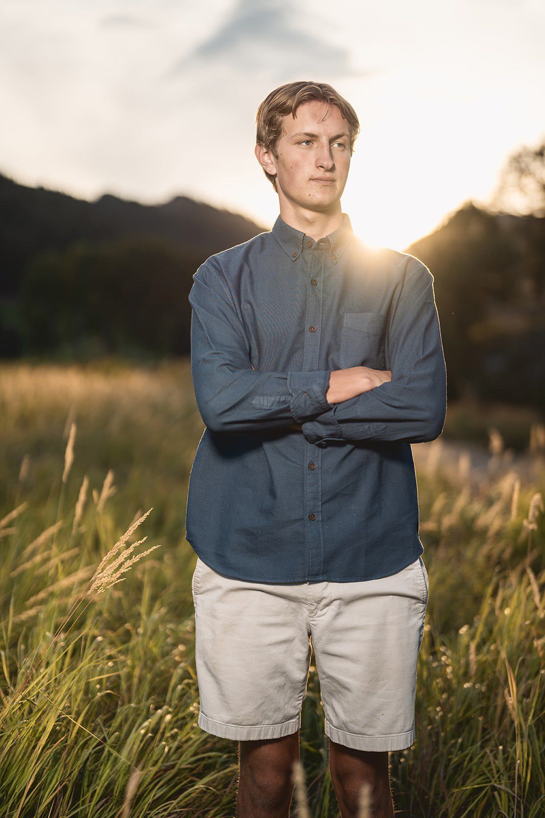 Young man in blue chambray shirt and white shorts standing in golden sunset field with arms crossed.