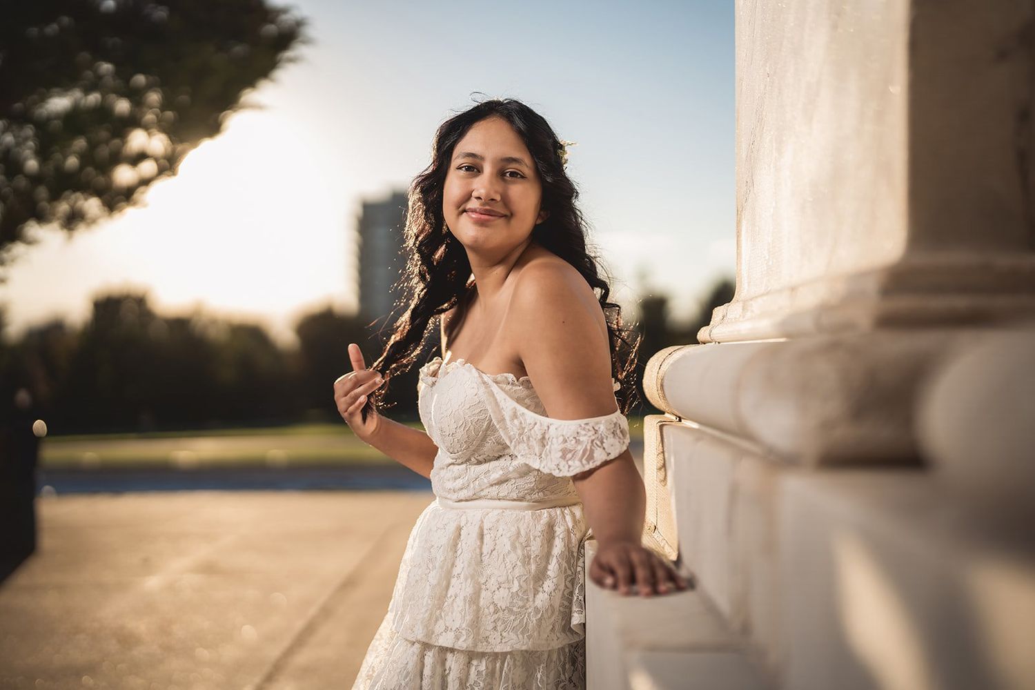 Young woman in white lace off-shoulder dress posing gracefully by stone columns at golden hour.