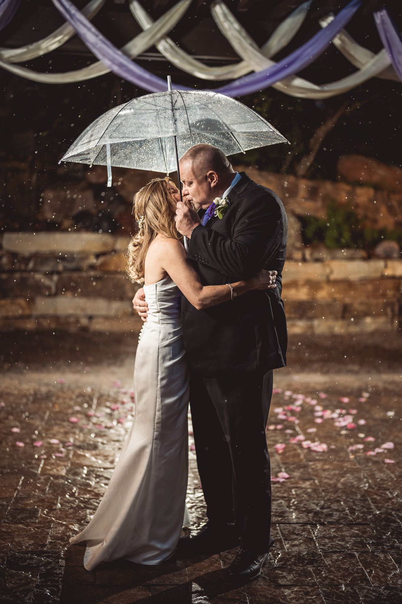 Bride and groom share a romantic kiss under a clear umbrella in the rain at their outdoor wedding ceremony.
