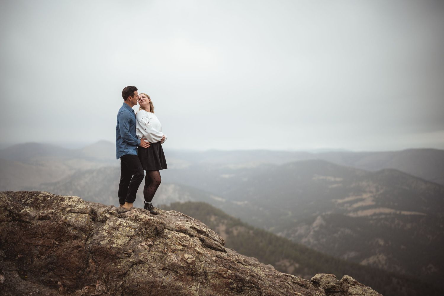 Couple embracing on mountain peak with dramatic mountain range vista in background.