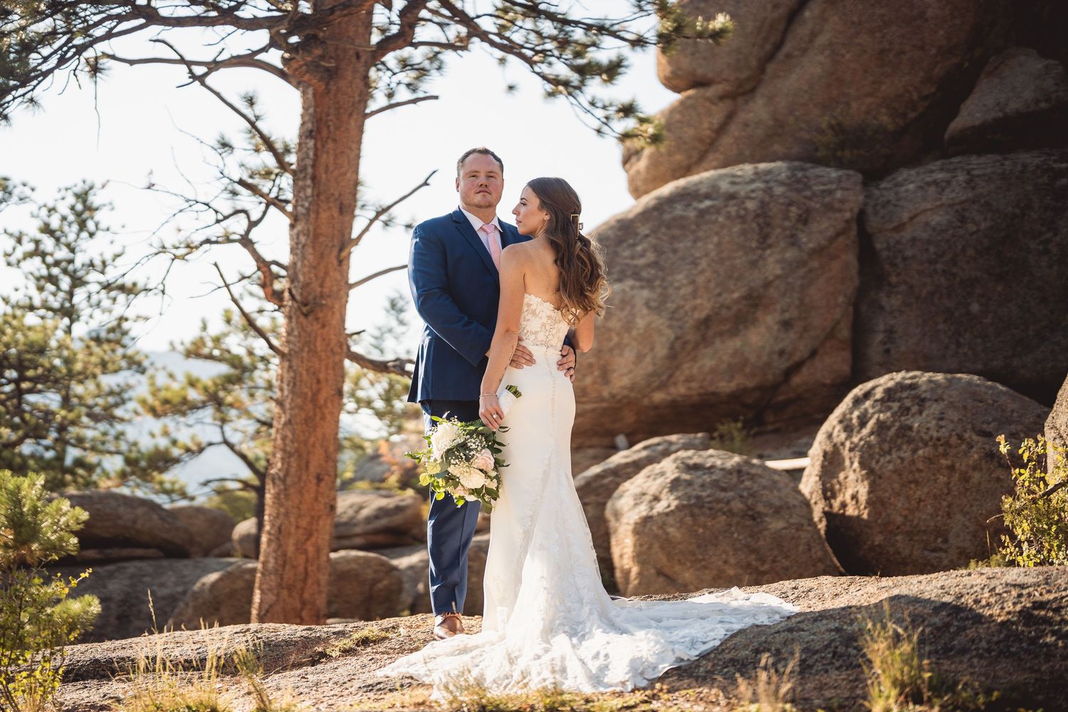 Wedding couple poses among large rocky outcroppings and pine trees in mountain setting.