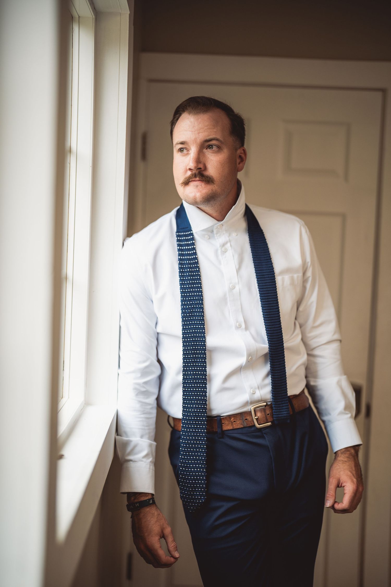 A man in formal attire adjusts his navy blue polka dot tie while getting dressed.