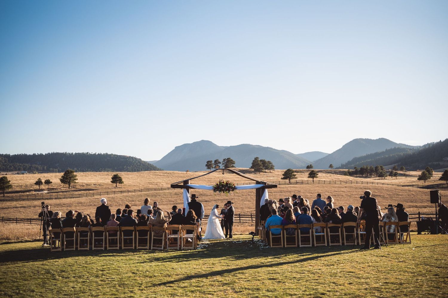 Panoramic view of outdoor wedding ceremony with mountains and guests seated on wooden chairs.