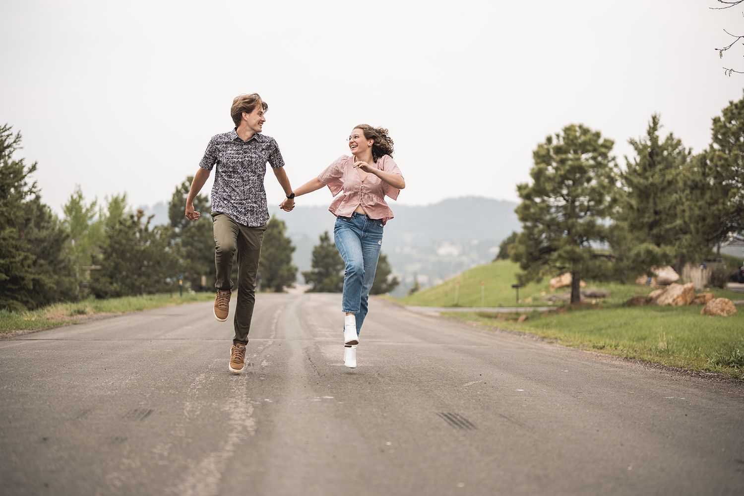 A young couple runs hand-in-hand down an open road surrounded by trees and mountains on a cloudy day.