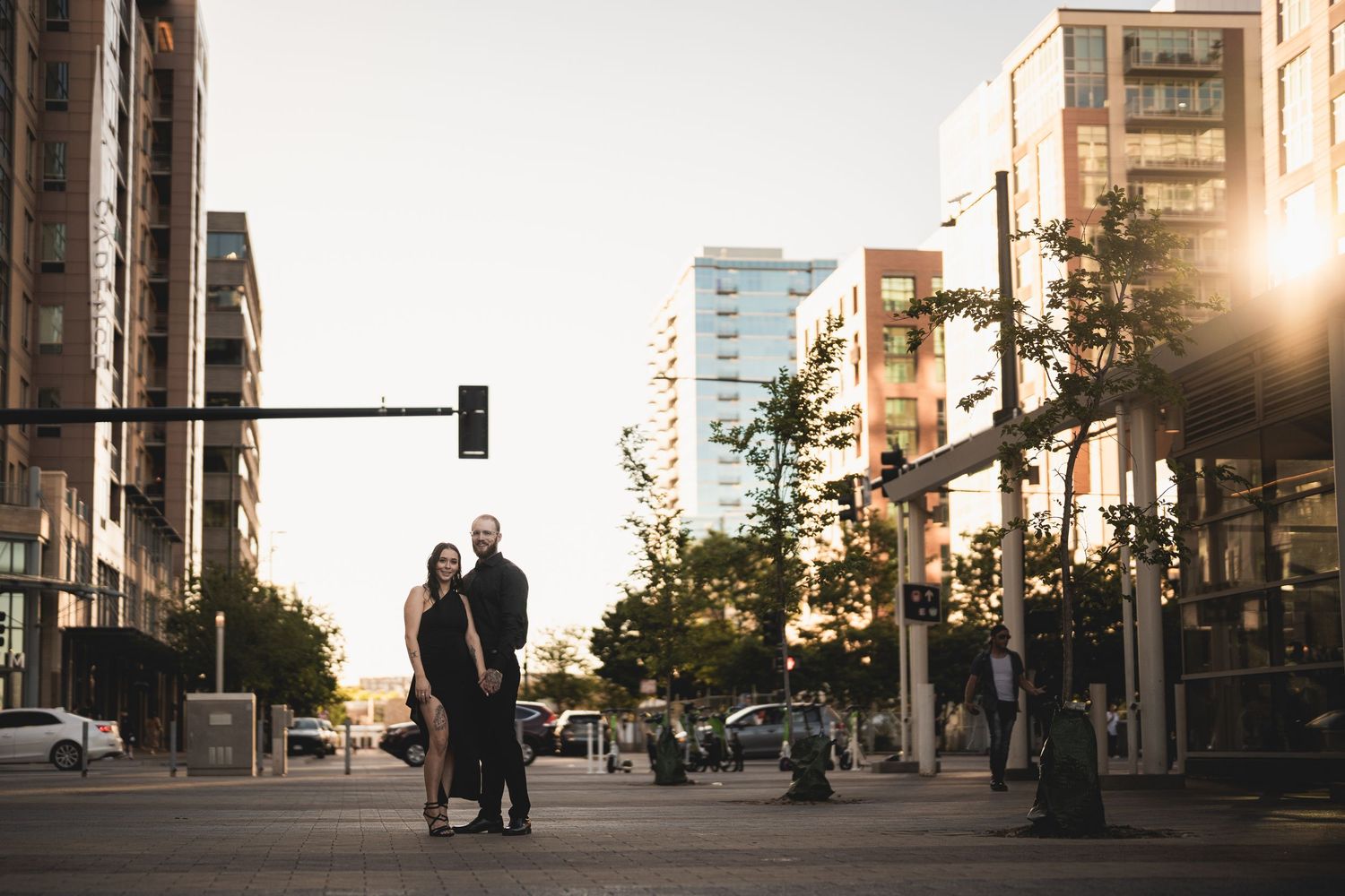 Couple standing on urban street corner at golden hour with city buildings and traffic light backdrop.