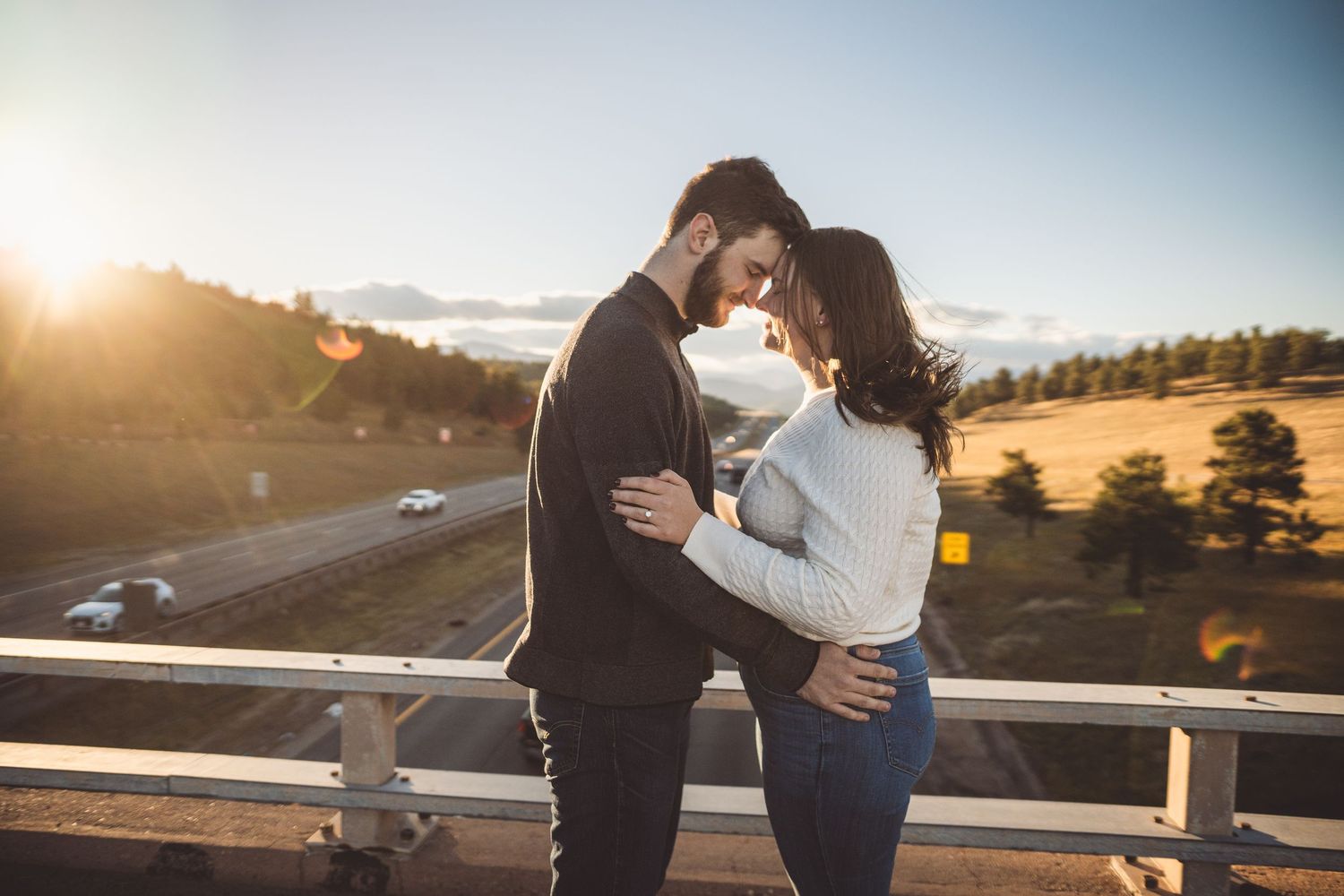 Romantic couple embracing and almost kissing on a bridge at golden hour sunset with highway and trees in background.