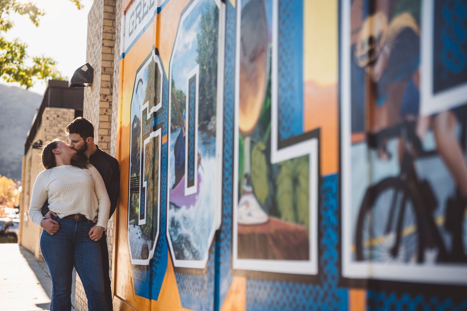 Couple sharing a romantic kiss in front of a vibrant colorful mural on a sunny day outdoors.