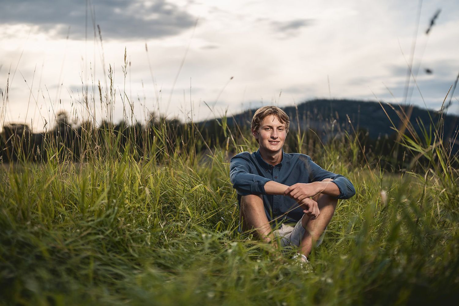 Young man sitting cross-legged in tall green grass with mountains visible in the background.