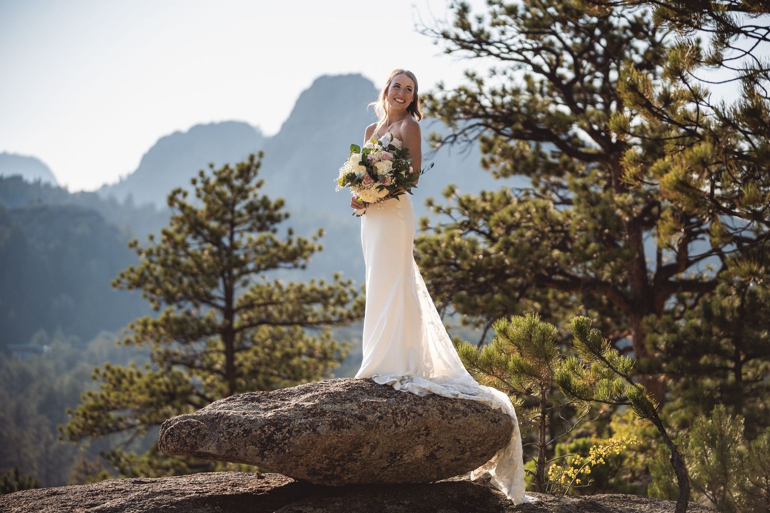 Bride in white gown holding bouquet stands on rock with mountain and pine tree backdrop.