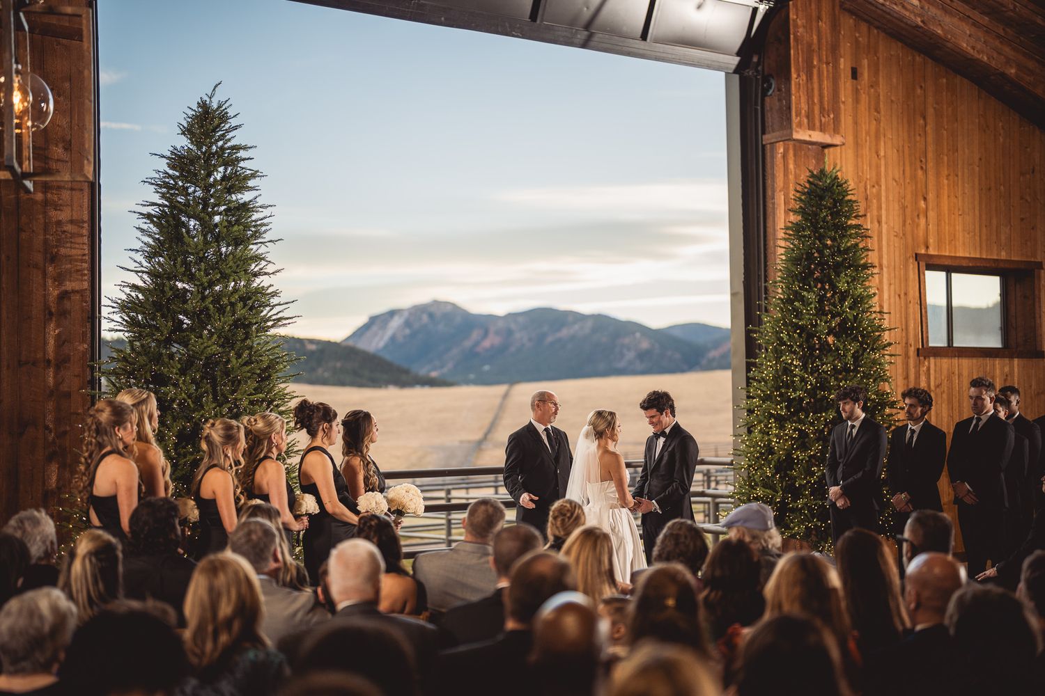 Wedding ceremony in rustic barn venue with mountain views, evergreen trees, bride and groom at altar with wedding party.