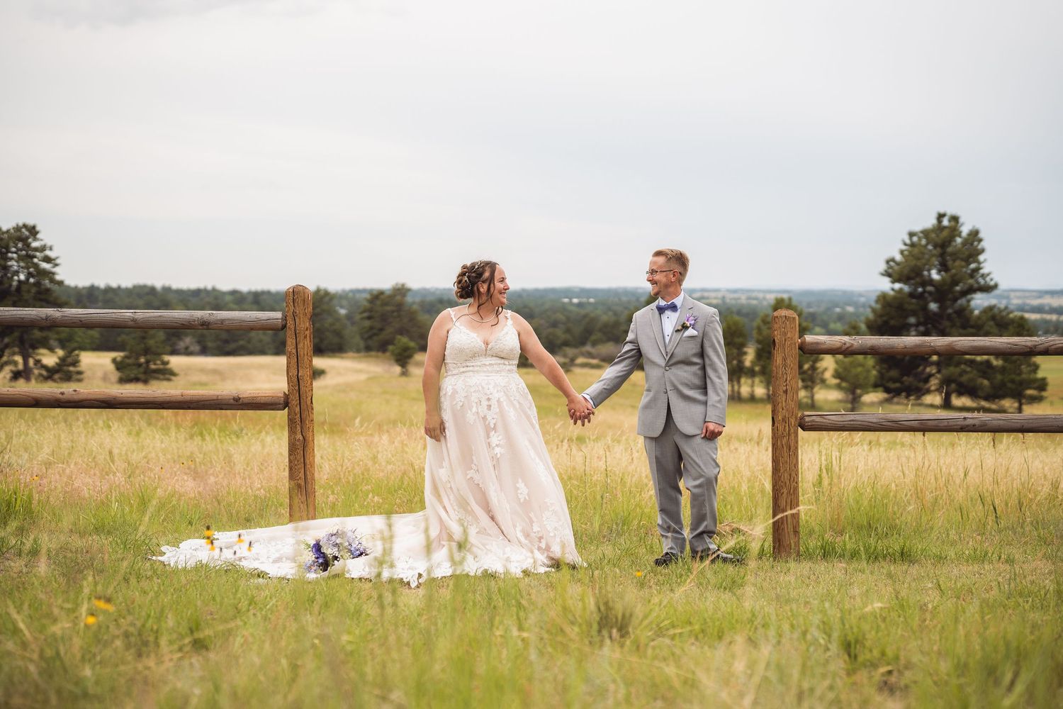 Wedding couple walks hand in hand through a sunny meadow with mountain views.