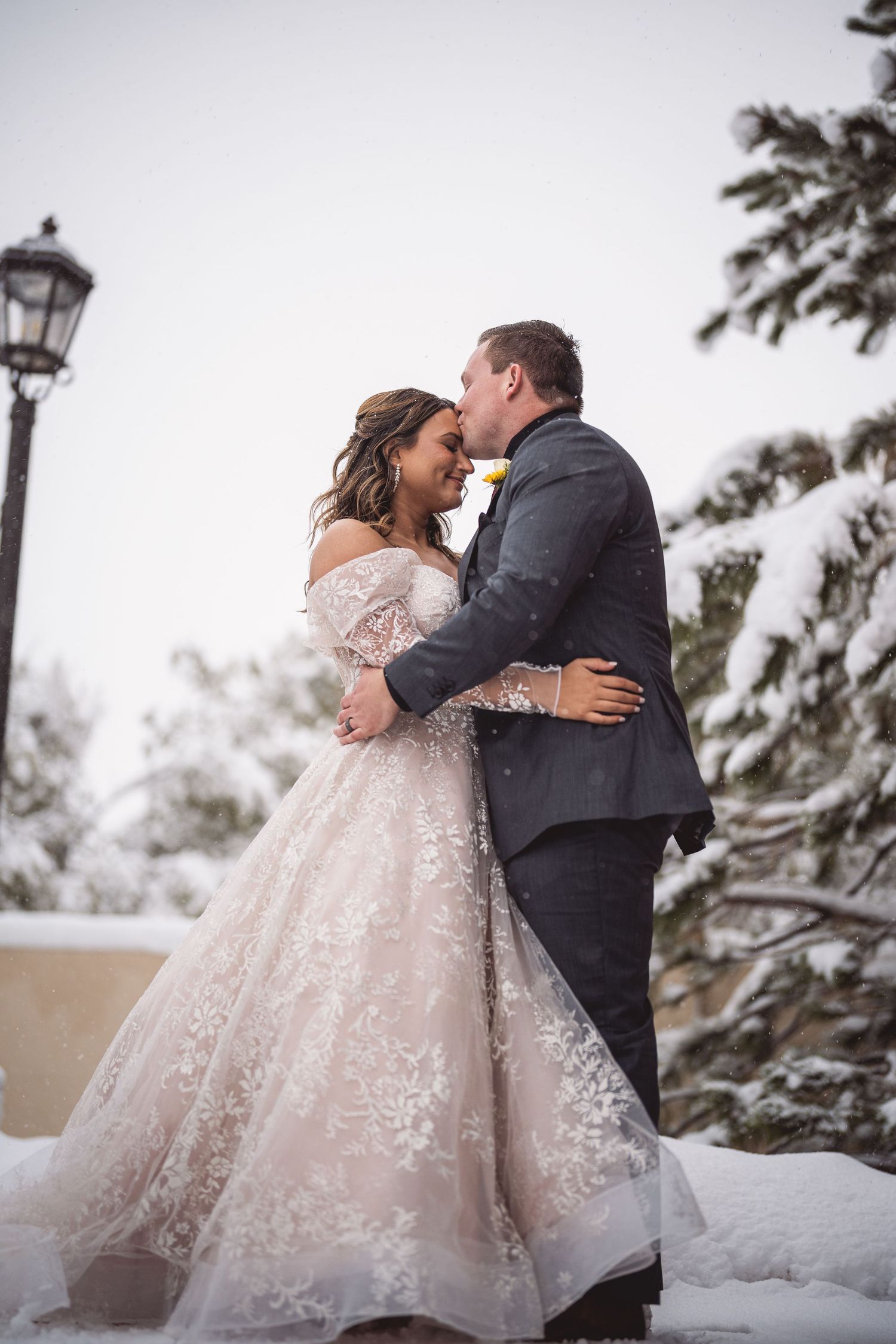 Newlyweds share intimate moments in a snowy winter landscape with snow-covered pine trees.
