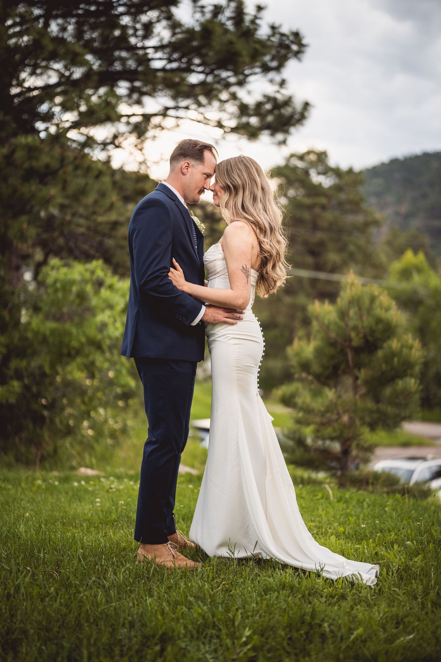Couple in wedding attire embracing on a grassy hillside with mountains in the distance.