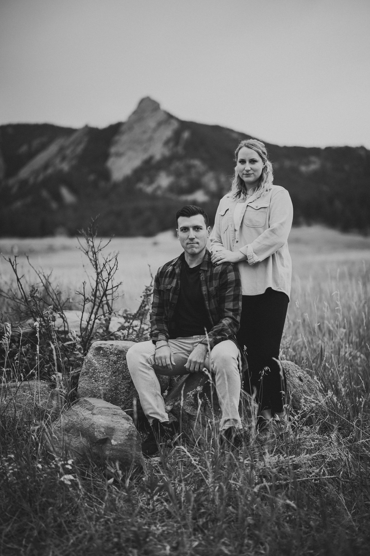 A black and white portrait of a couple sitting together in a grassy mountain meadow.