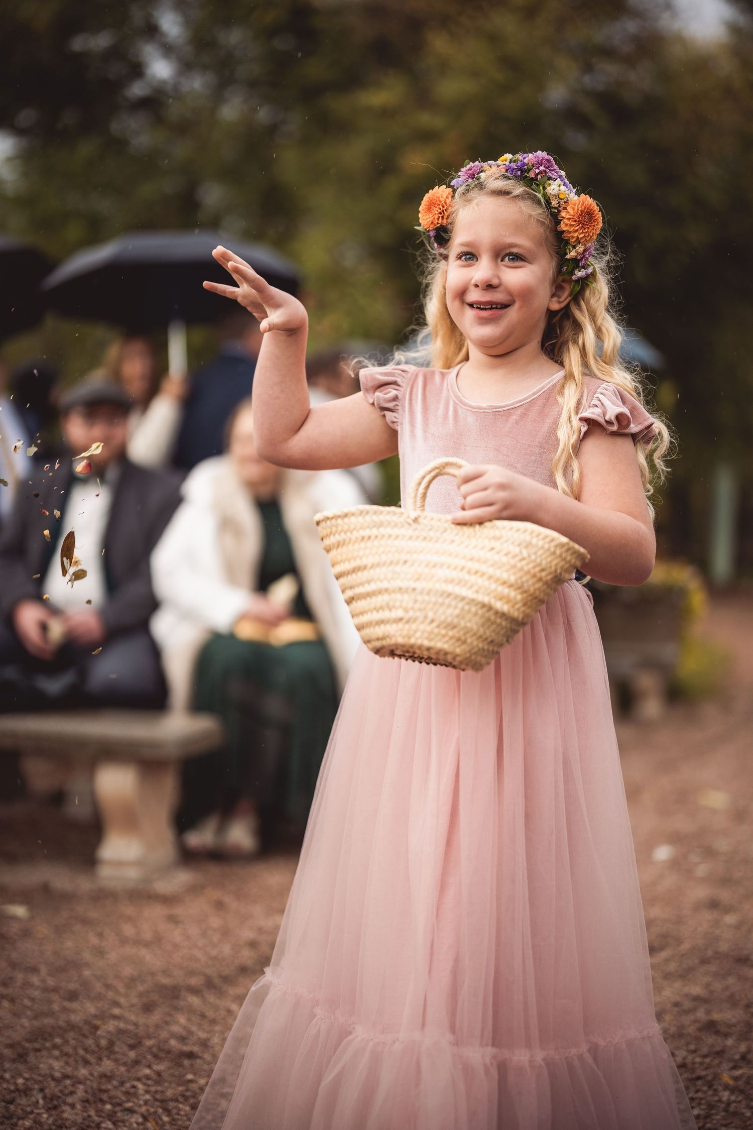 Flower girl in pink dress carries basket while walking down gravel path at outdoor wedding.