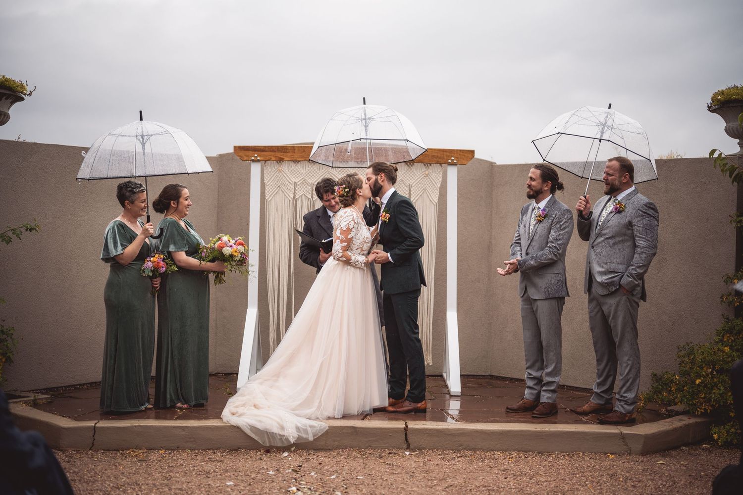 Outdoor wedding ceremony with guests holding umbrellas against white walls.
