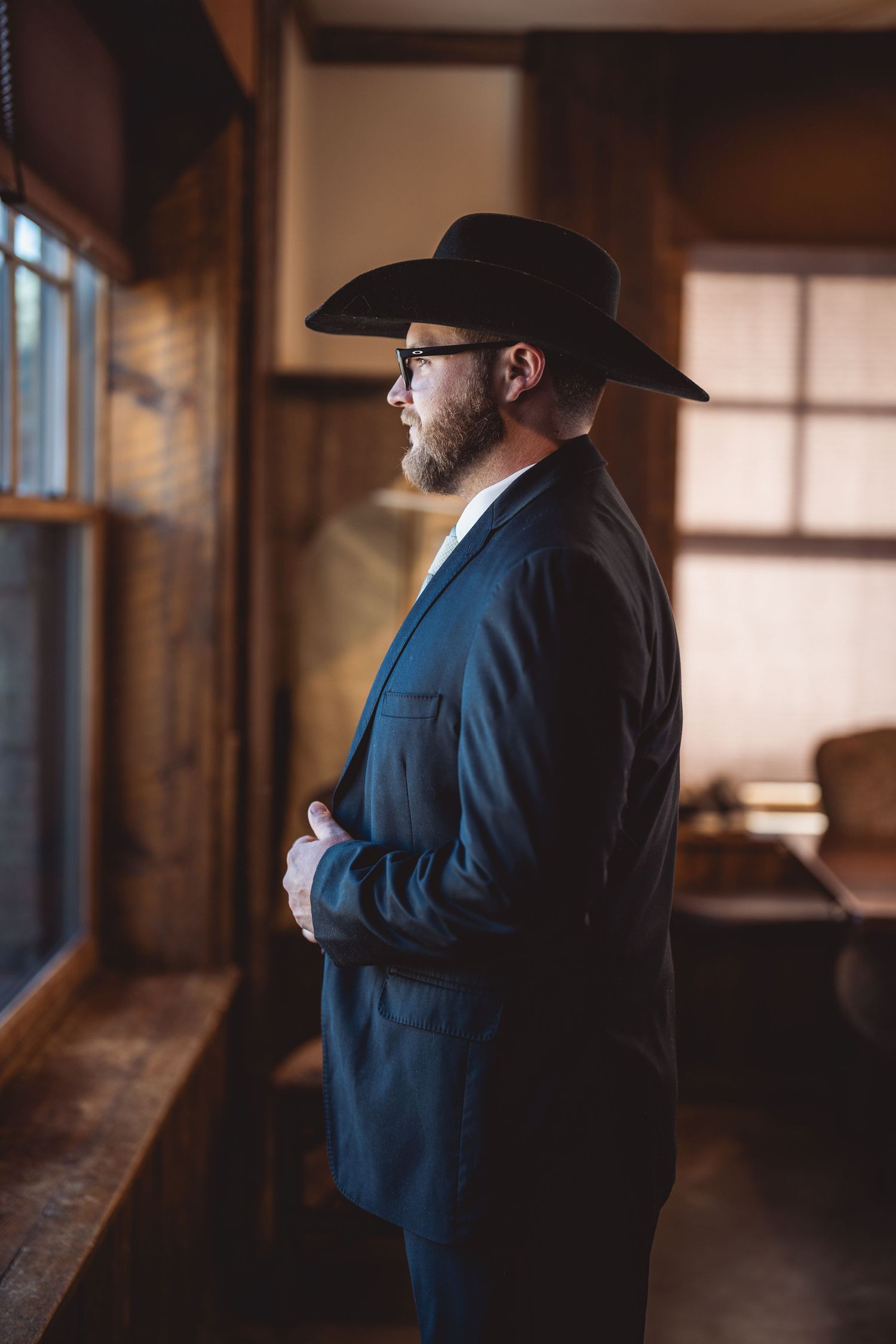 A silhouetted figure in a cowboy hat and suit stands in dramatic window lighting in a rustic wooden room setting.