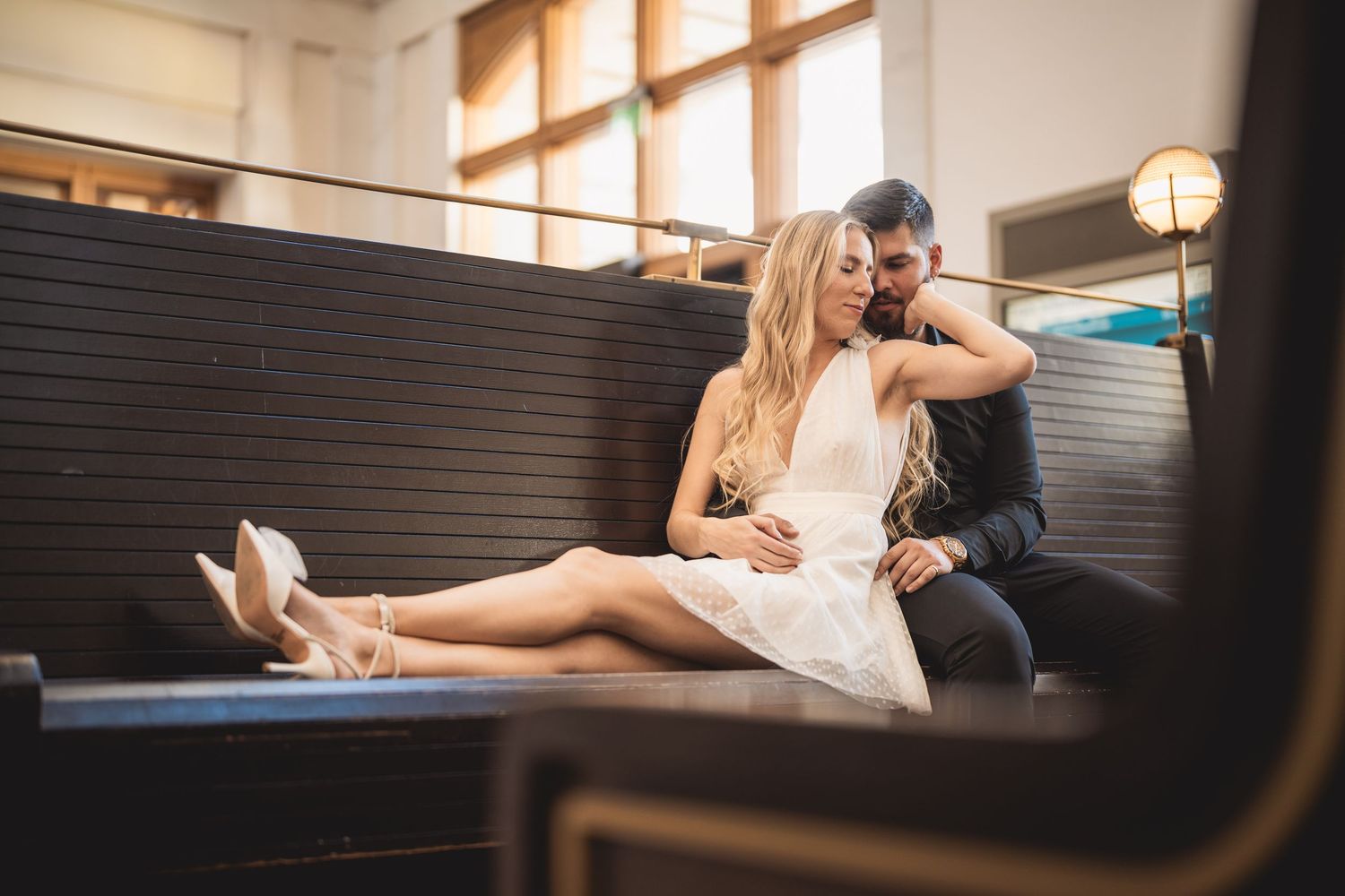 A couple in wedding attire sits intimately on a dark bench in an elegant indoor venue.