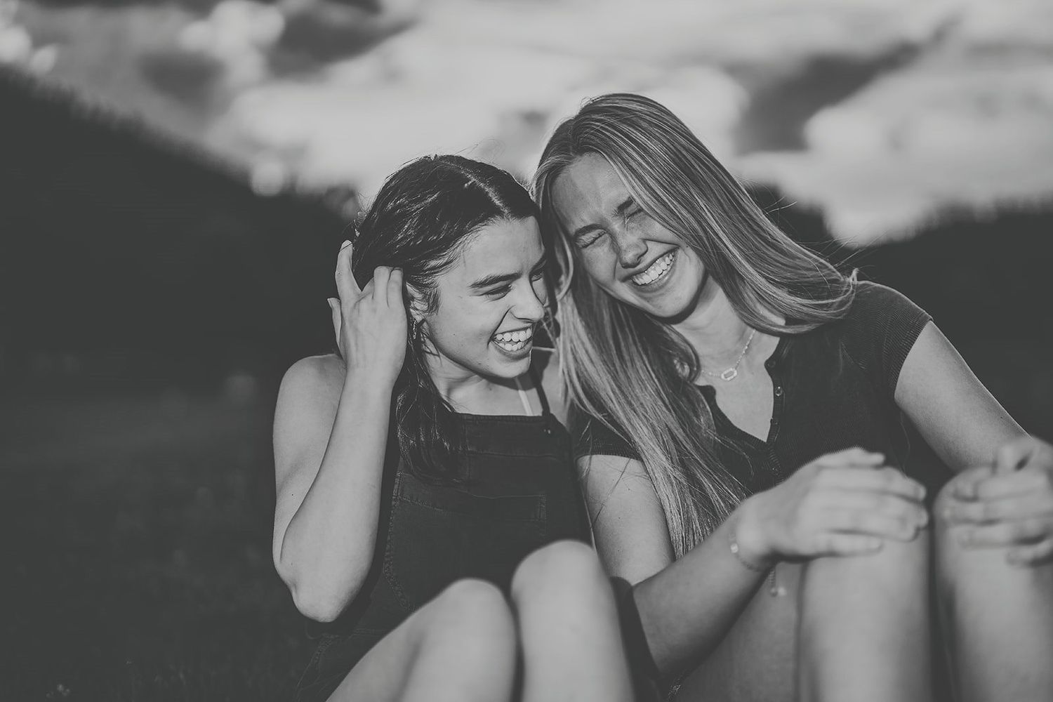 Two young women laughing together outdoors in black and white photo, sitting close and sharing a joyful moment.