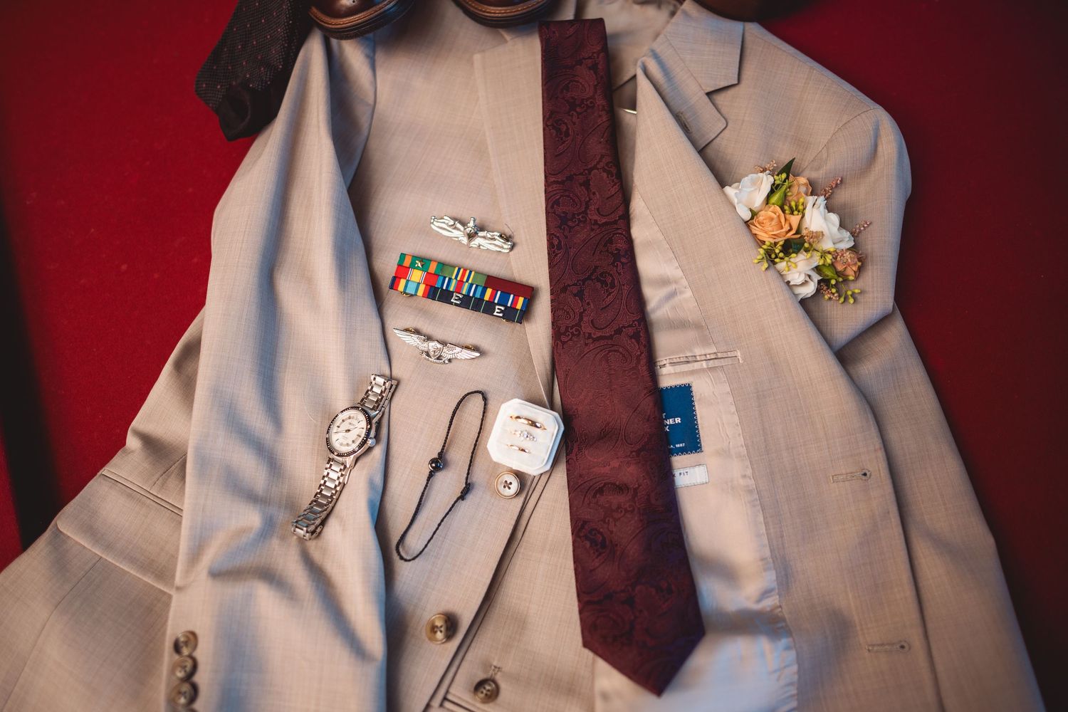 Wedding suit details with burgundy tie and lapel pins against red background.