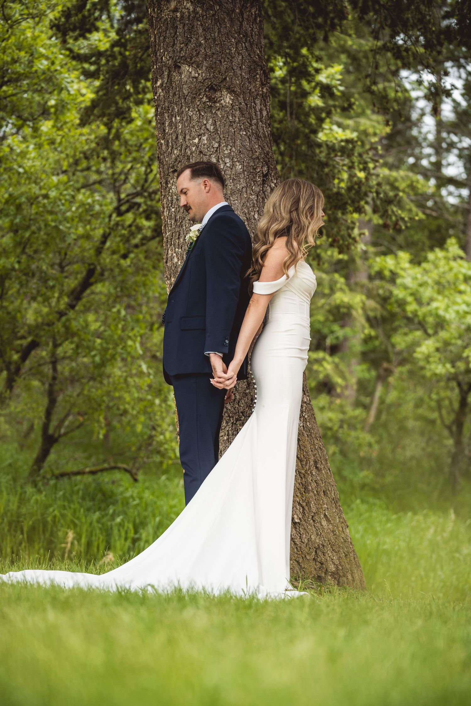 Romantic wedding couple holding hands behind a large tree trunk in a lush green field.