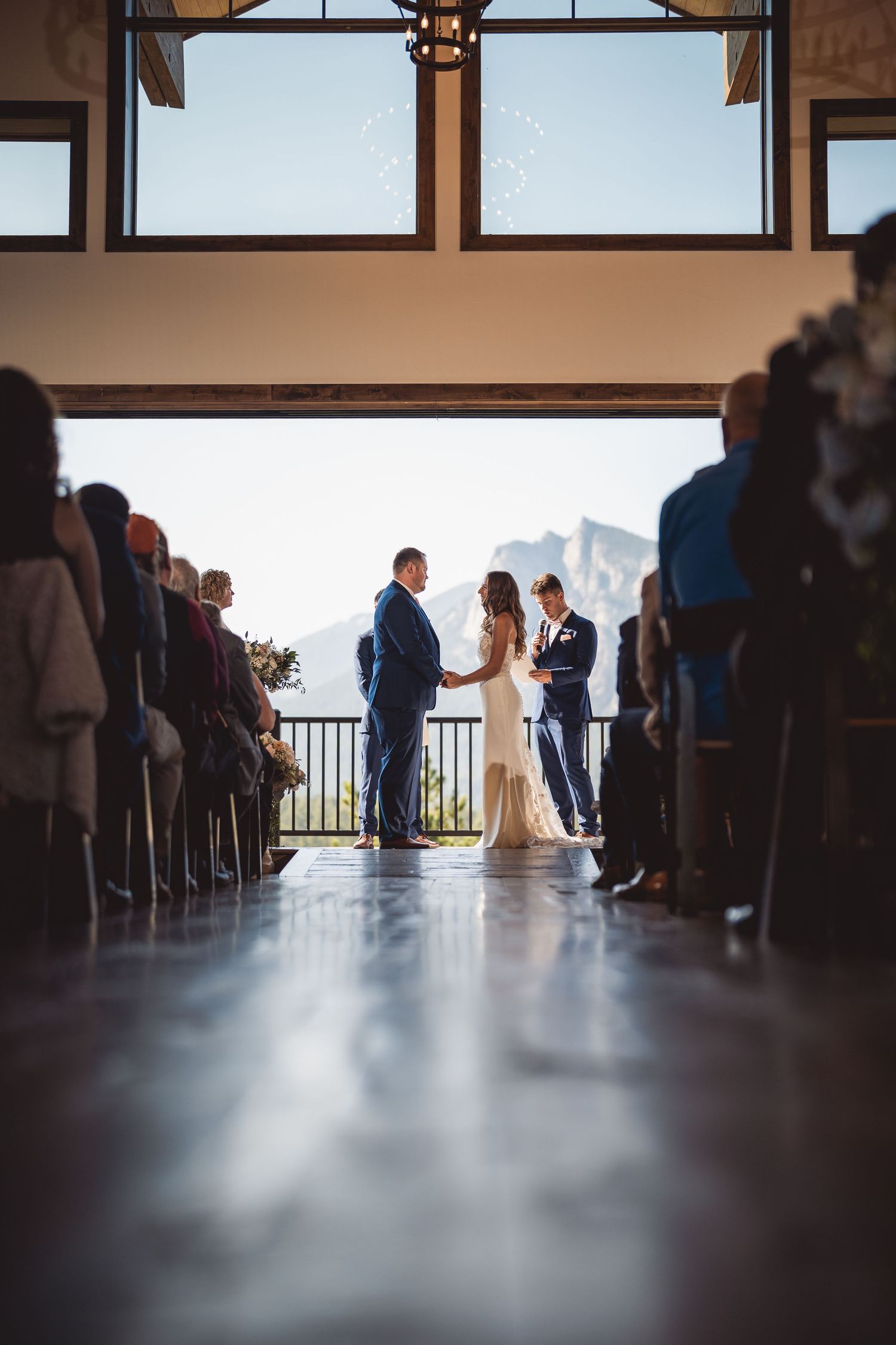 Wedding guests gather in a venue with silhouettes against large windows overlooking mountains.