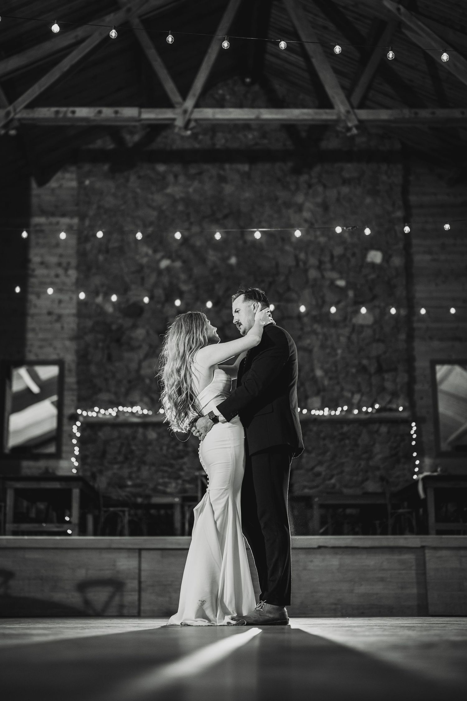 Black and white photo of dancing silhouettes in a rustic barn with twinkling lights overhead.