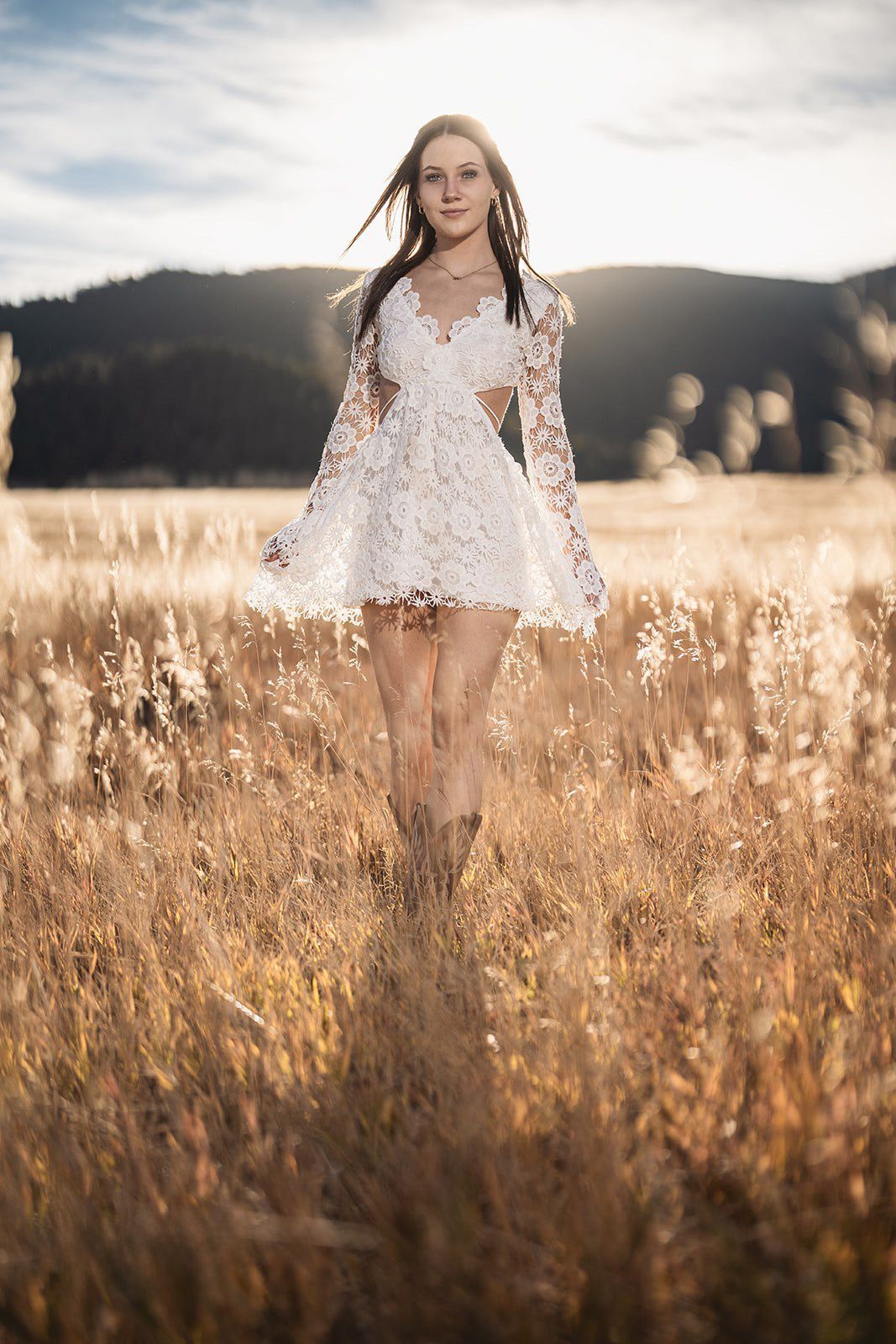 Young woman in white lace dress and cowboy boots walking through golden sunlit field with mountains.
