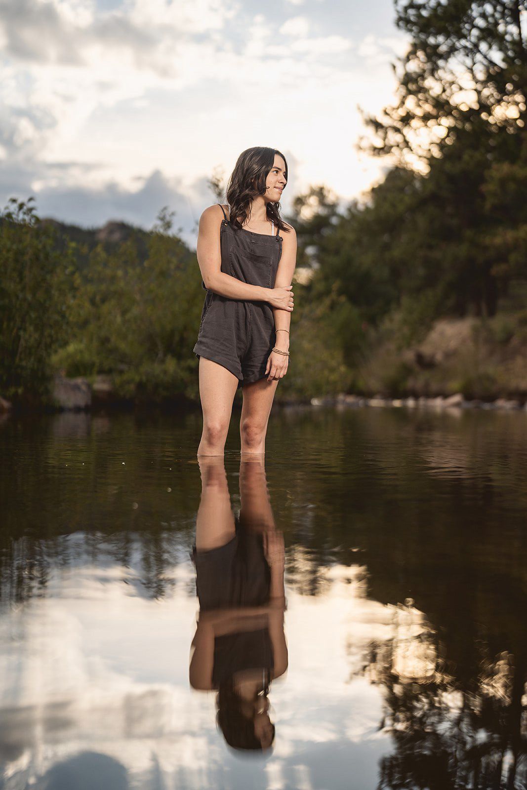 Young woman in dark romper standing in calm reflective water at sunset with lush green trees in background.