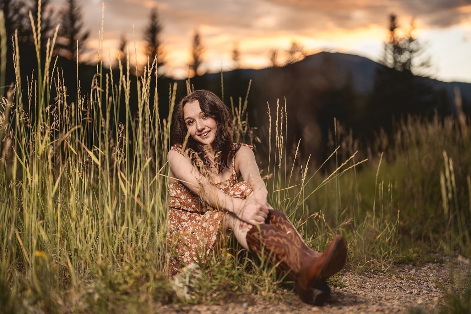 Laughing young woman in floral dress and cowboy boots sitting in tall grass at mountain sunset.
