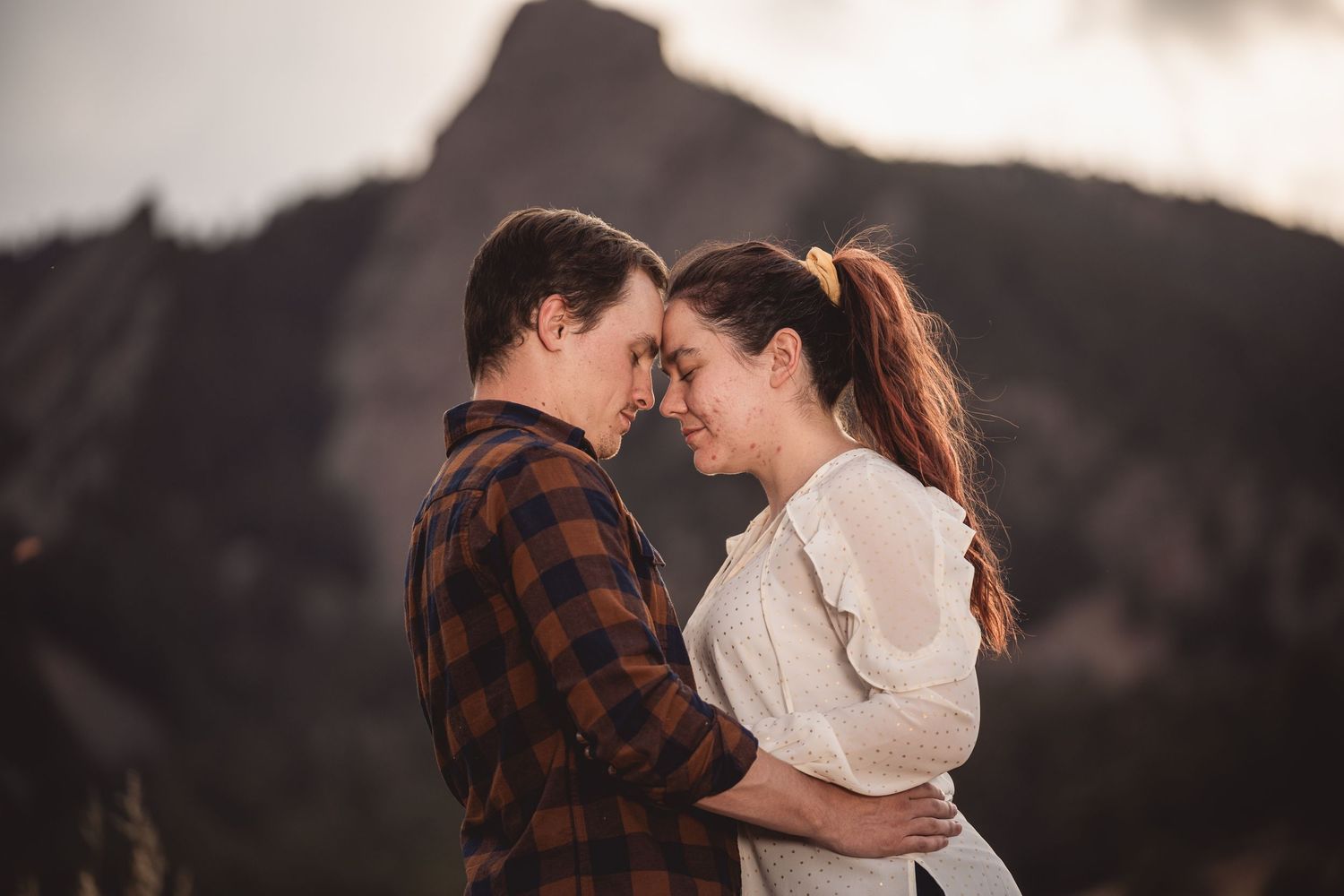 Couple sharing a romantic forehead touch with mountains in the background during golden hour outdoor engagement session.