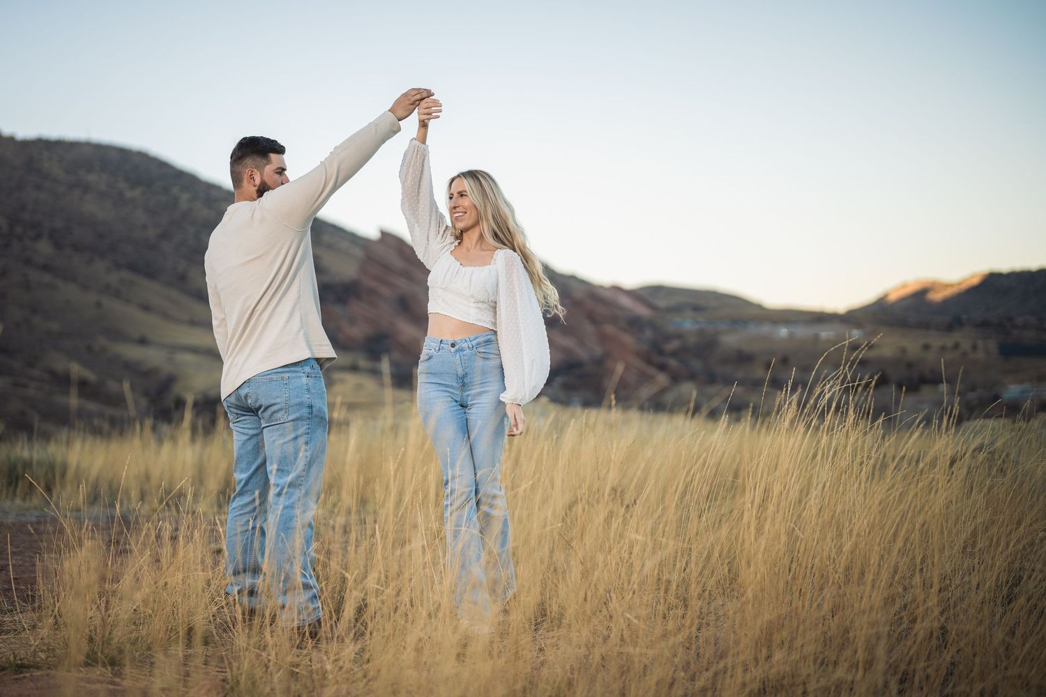 Couple dancing in golden field with mountain backdrop, wearing casual white tops and jeans on a sunny day.
