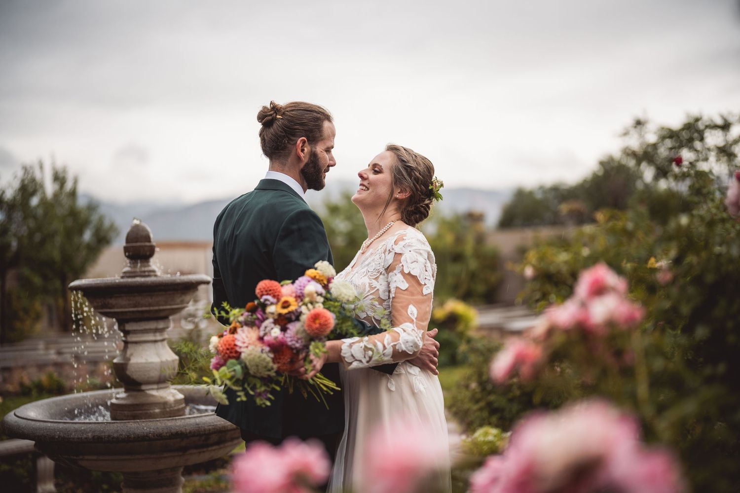 Romantic garden wedding portrait with couple surrounded by pink roses and stone architecture.