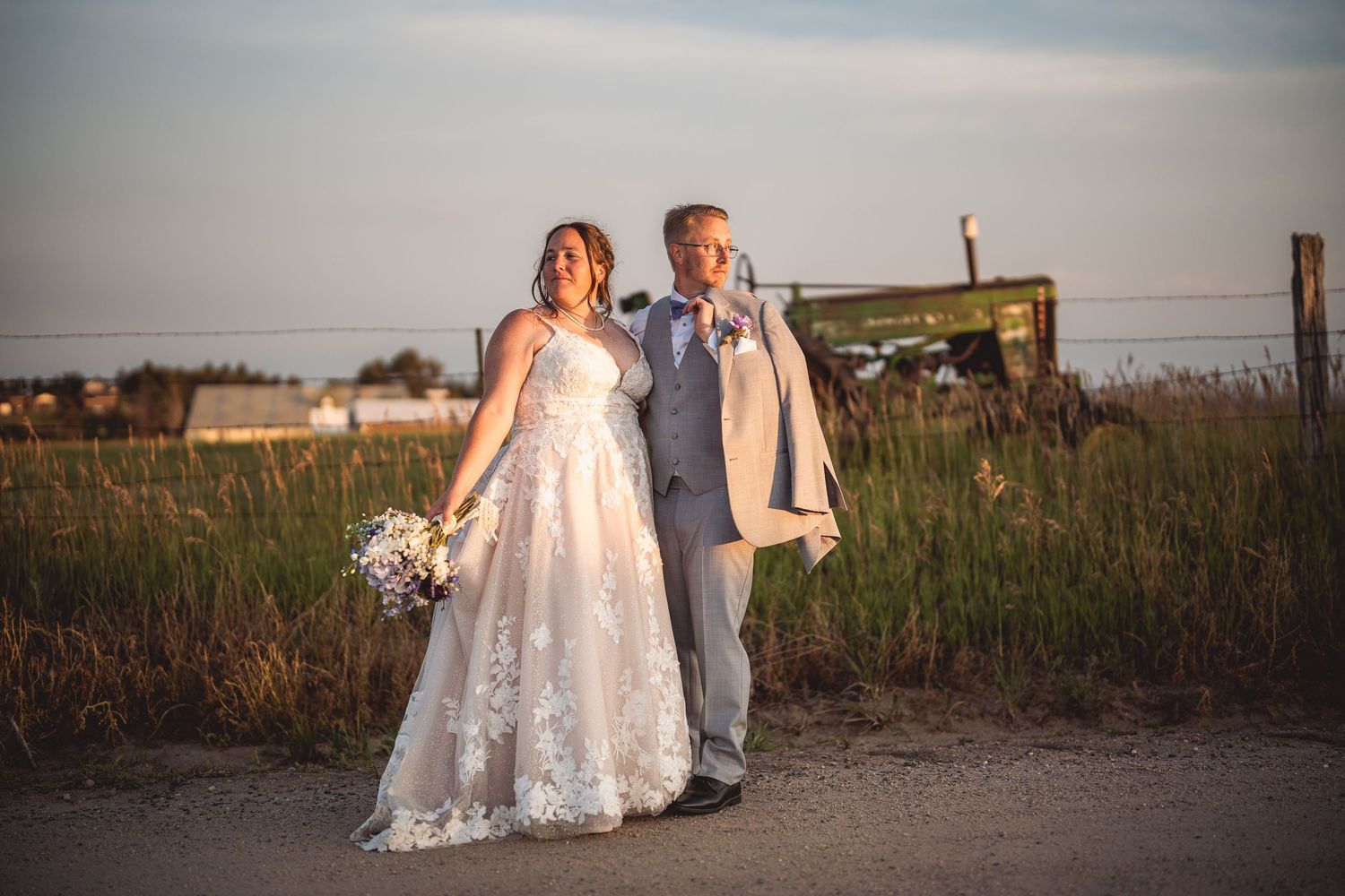 Couple stands together on rural path during golden hour with reception venue in distance.
