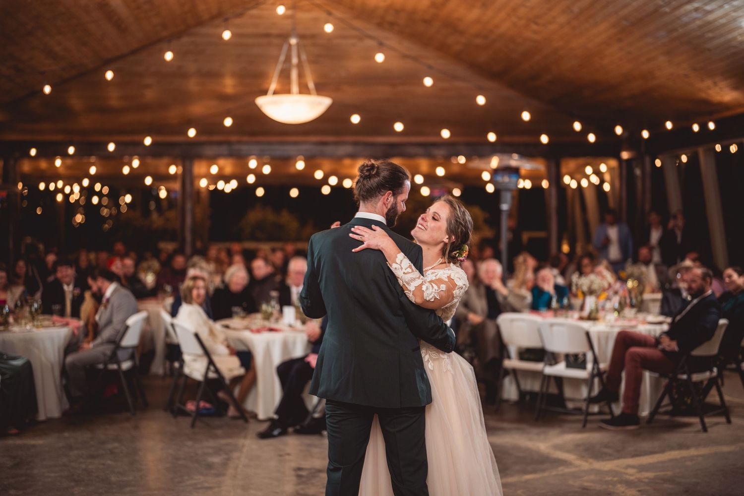 First dance at wedding reception under wooden beams and string lights with guests watching.