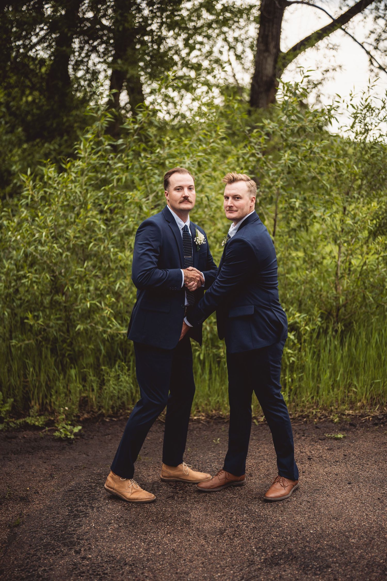 Two groomsmen in navy suits performing playful dance moves on dirt path.