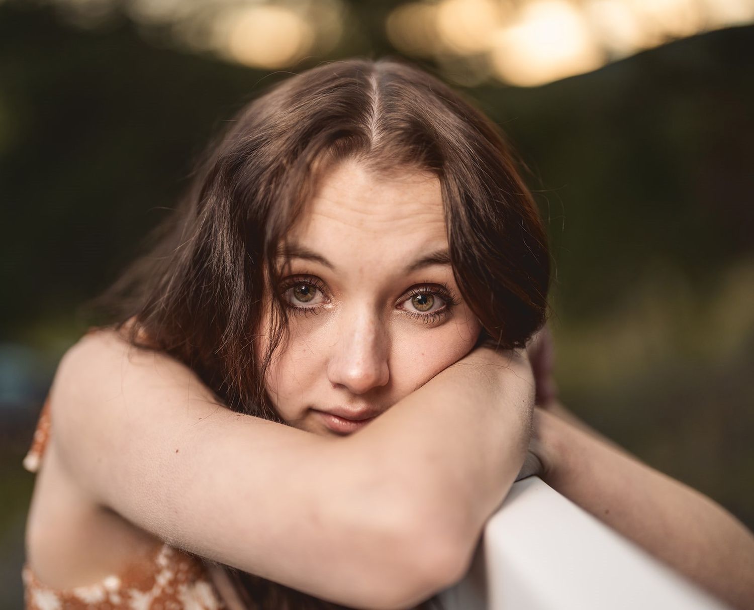 Close-up portrait of young brunette woman with green eyes resting chin on arms in soft light.