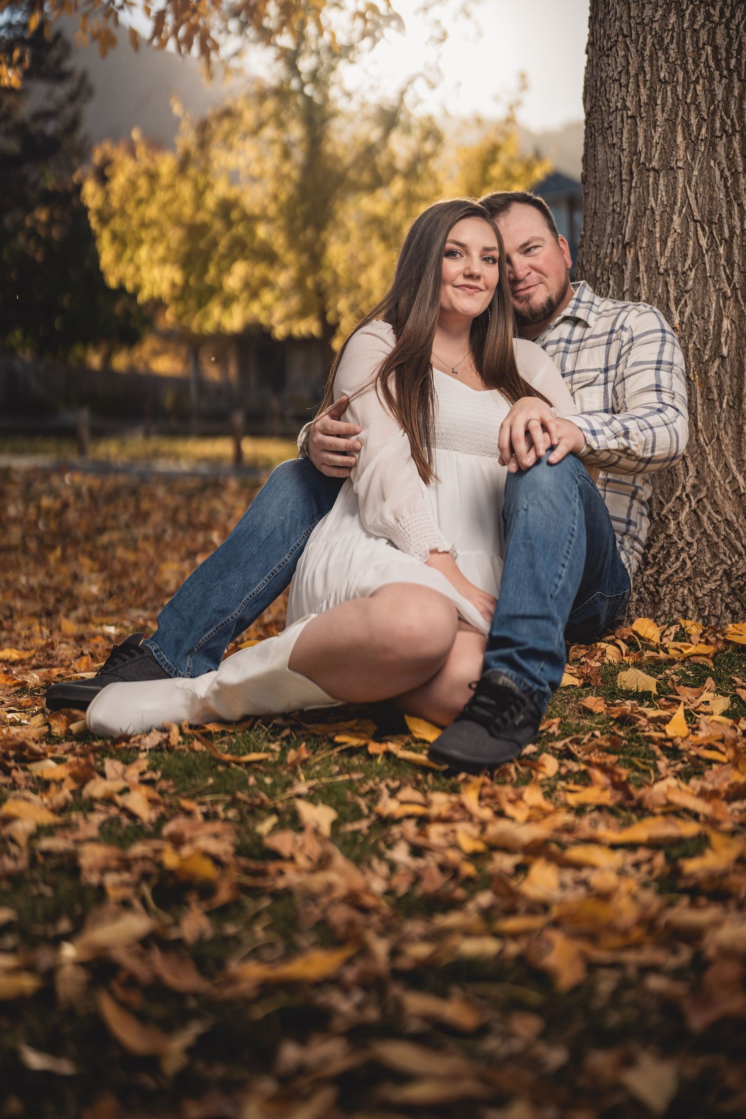 Couple sitting together against a tree surrounded by golden autumn leaves in a warm fall outdoor setting.