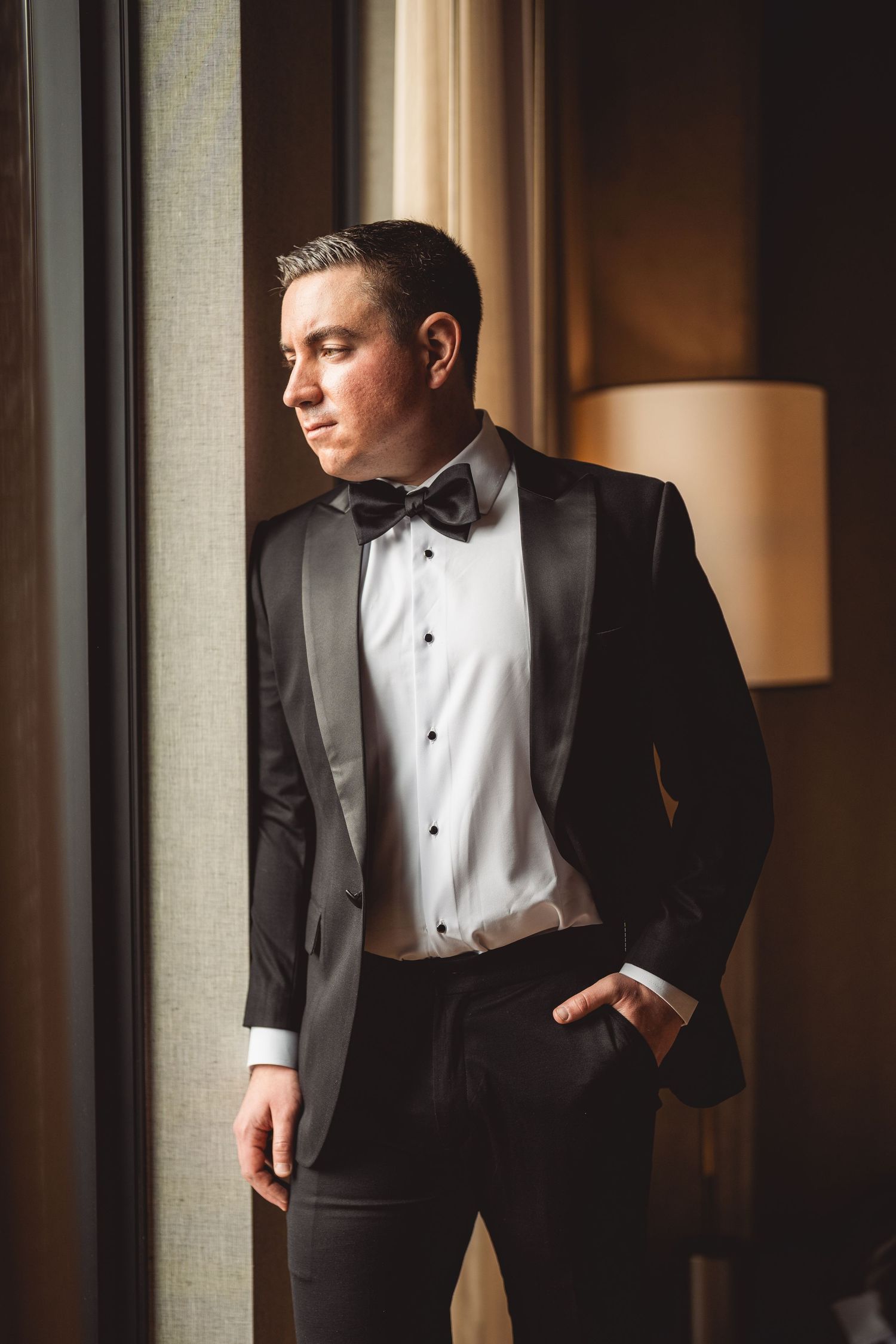 Groom in black tuxedo with bow tie poses by window with natural lighting.