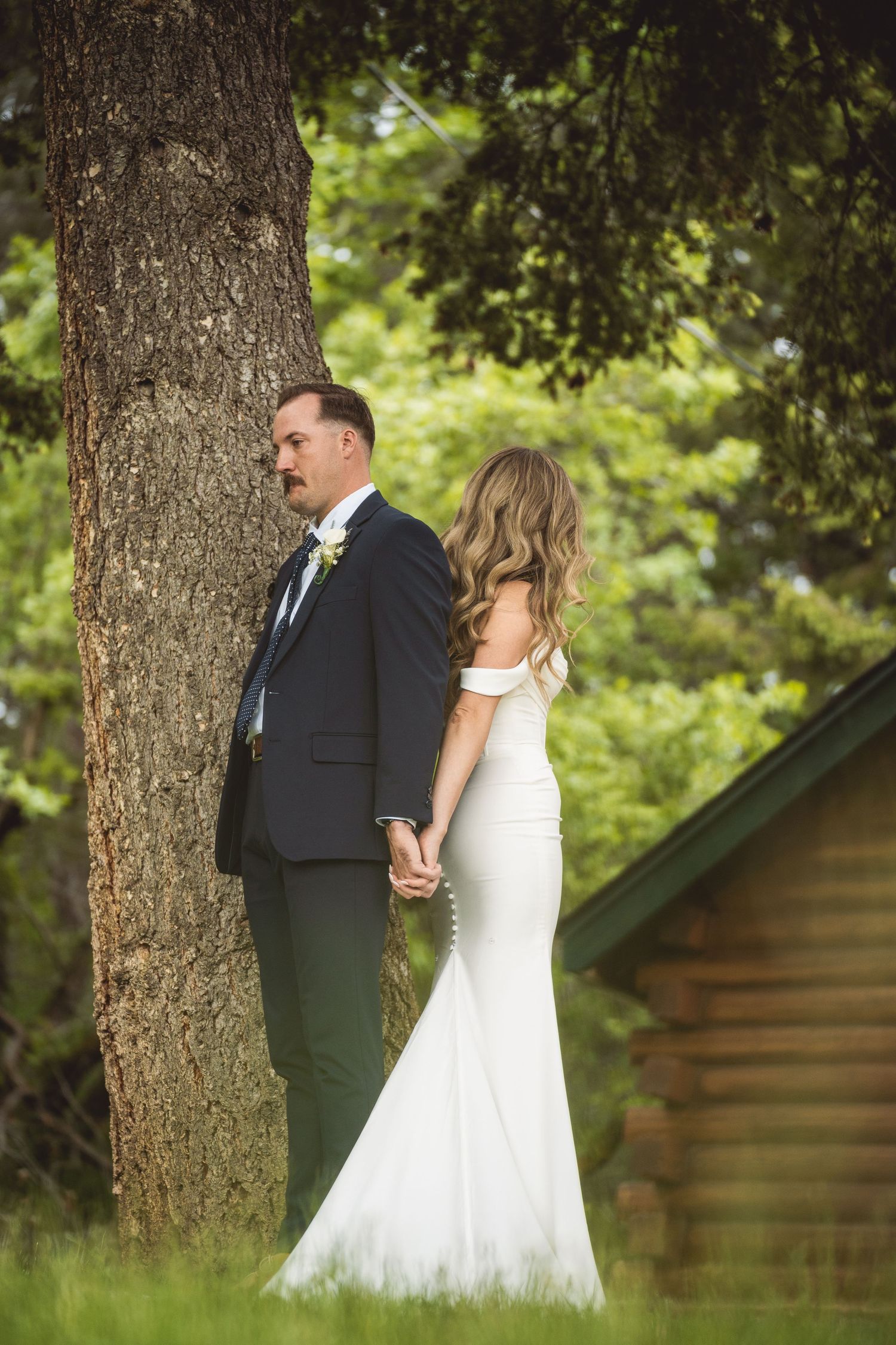 Wedding couple posing together under large tree with rustic cabin in background.