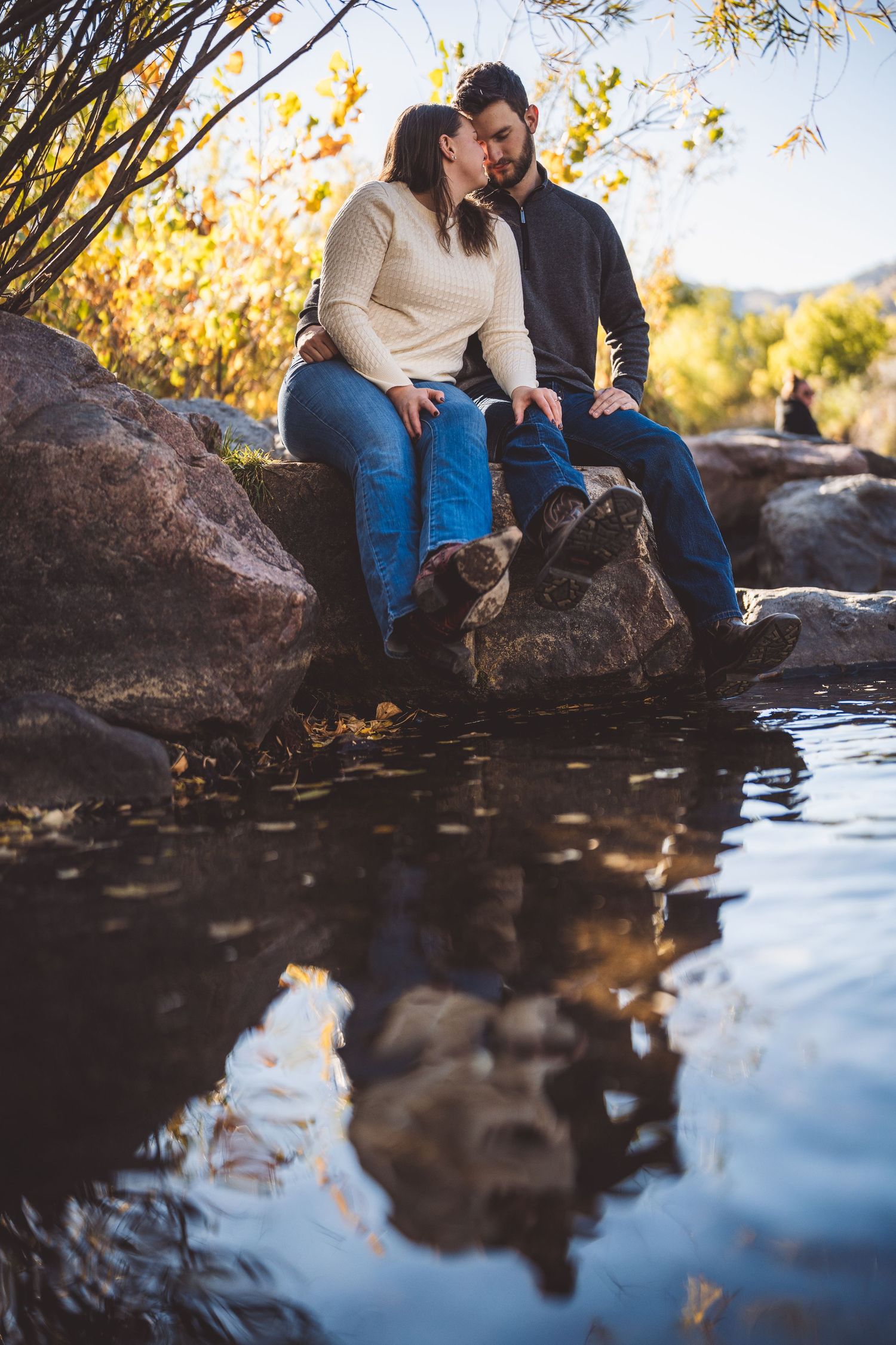 Couple sits by reflective water with autumn foliage creating beautiful nature setting.