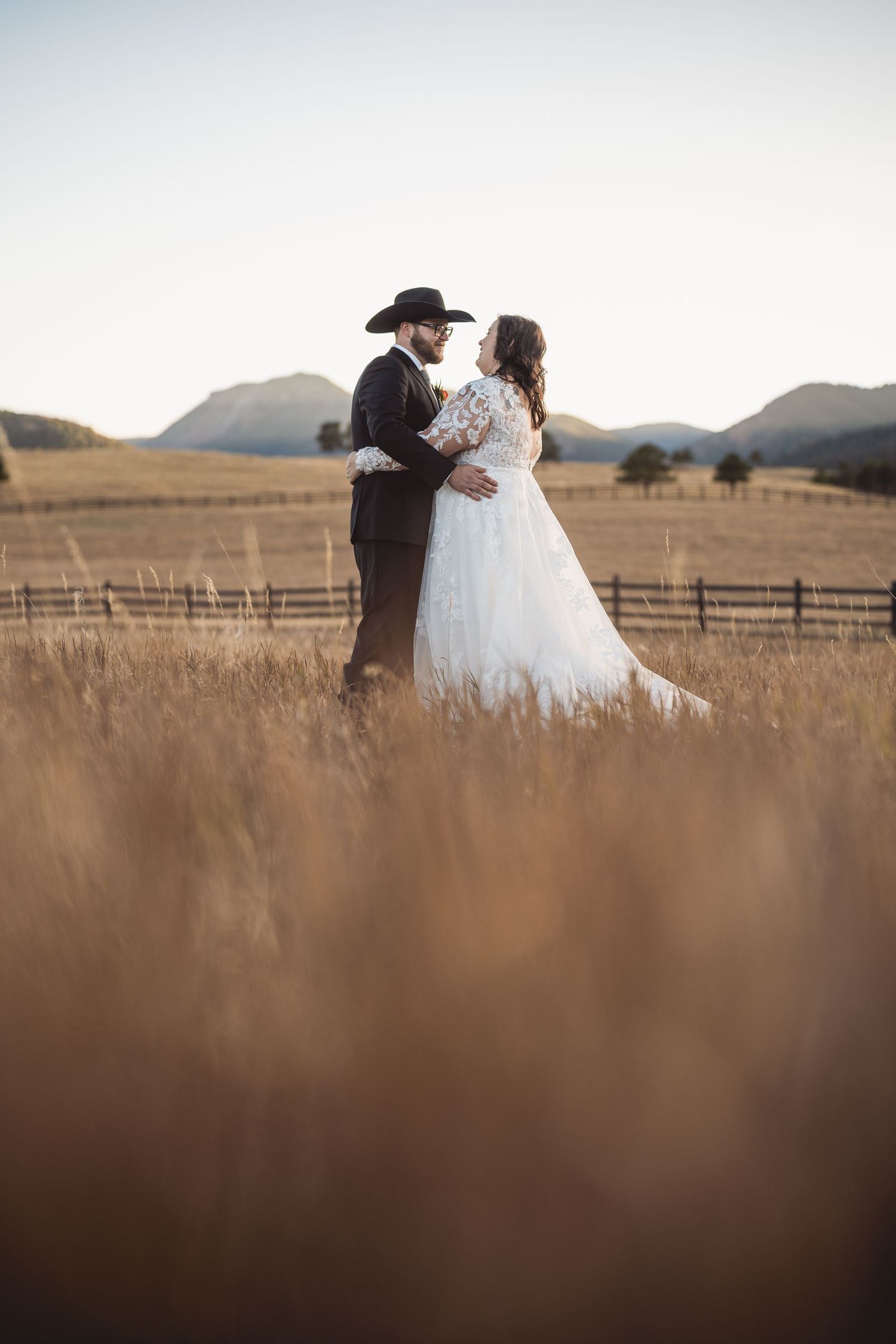 Romantic couple in wedding attire silhouetted against rustic mountain landscape and wooden fencing.