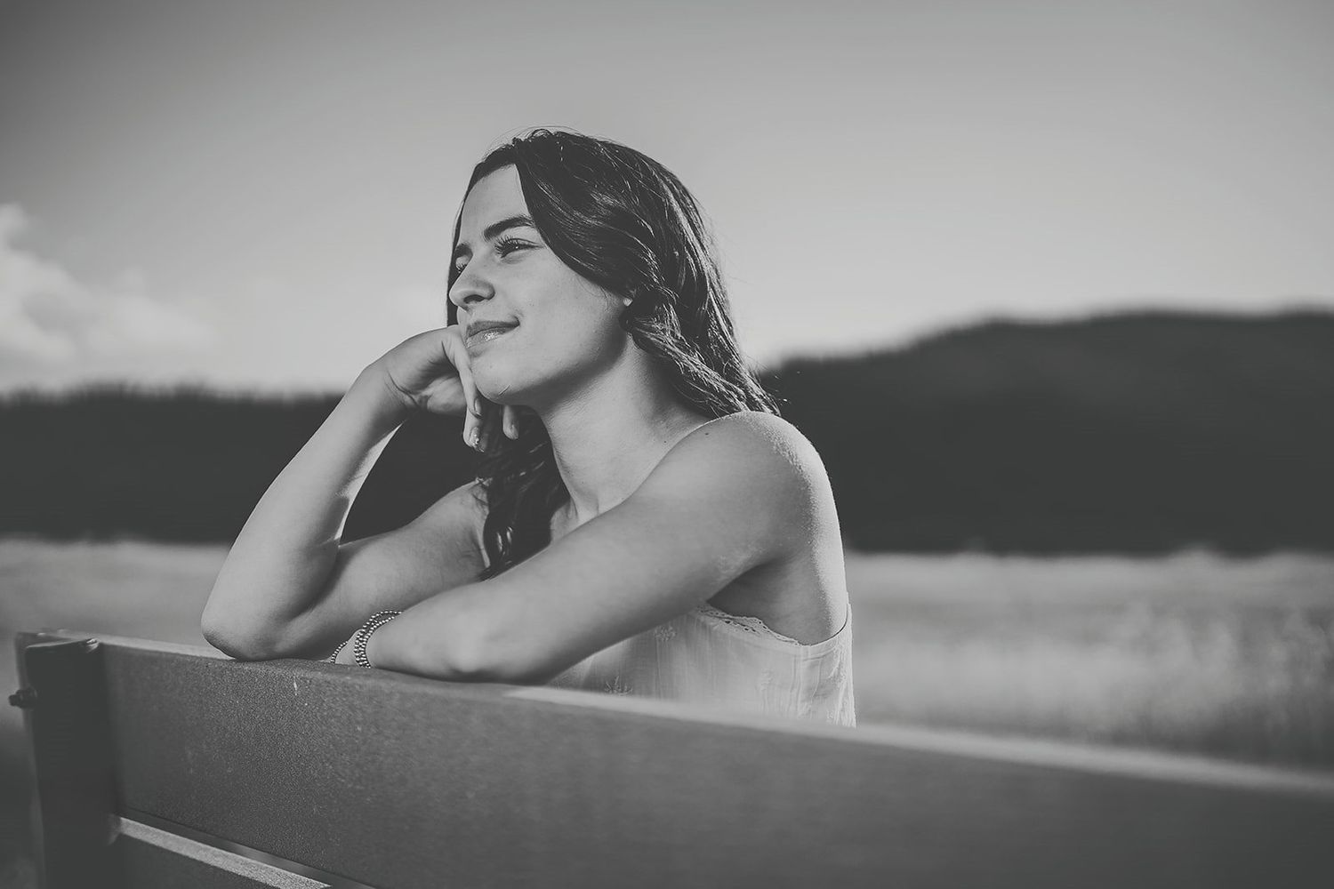 Black and white portrait of a young woman sitting on a bench outdoors, smiling and gazing into the distance.