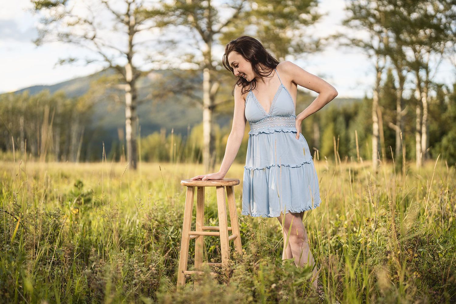 Young woman in light blue dress standing beside wooden stool in sunny meadow.