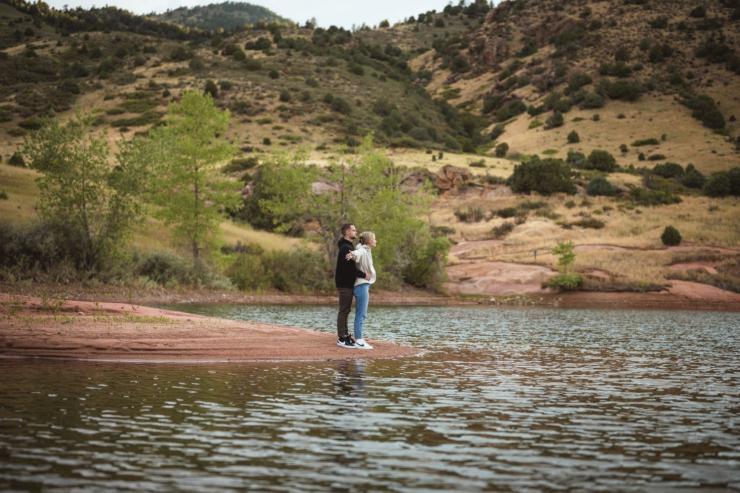 Couple embracing on a rocky shoreline beside a calm lake with rolling green hills in the background.