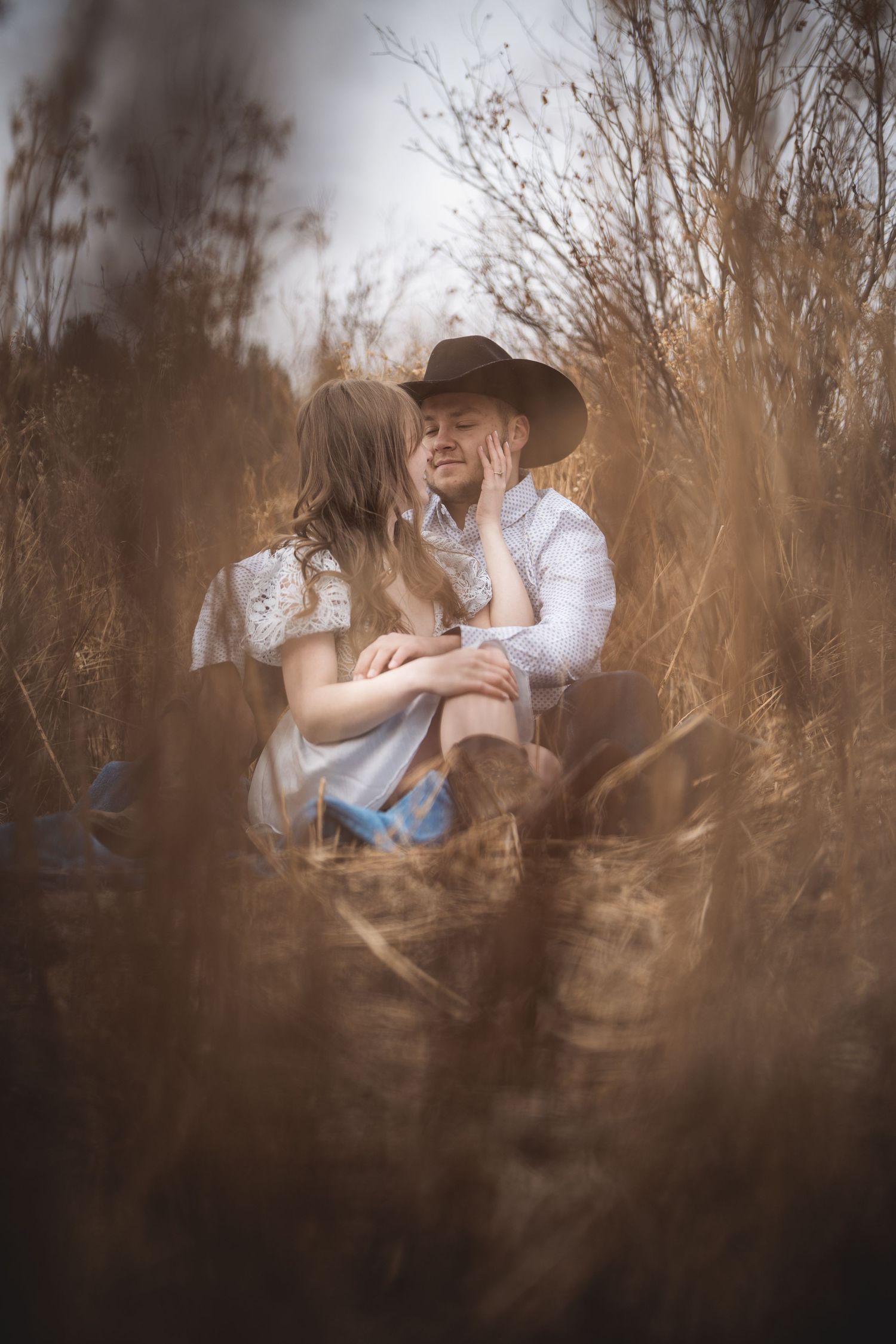 A romantic couple sits among tall dried grass during a dreamy outdoor photoshoot.