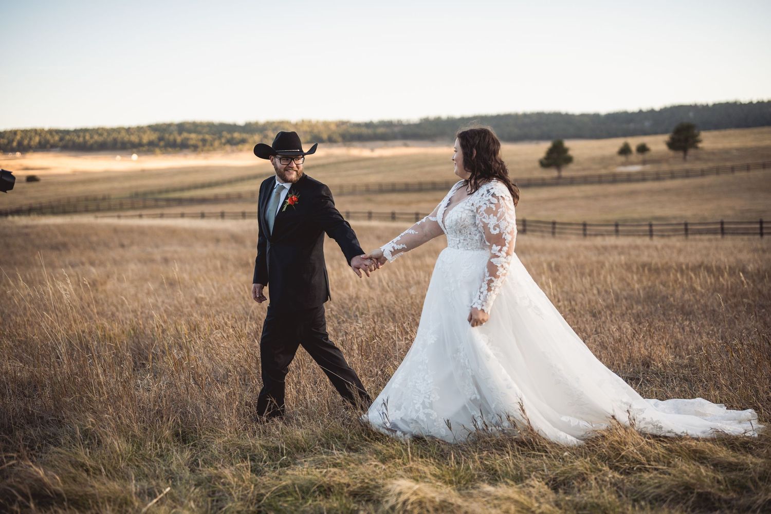 Couple in wedding attire walking through golden prairie grass at sunset.