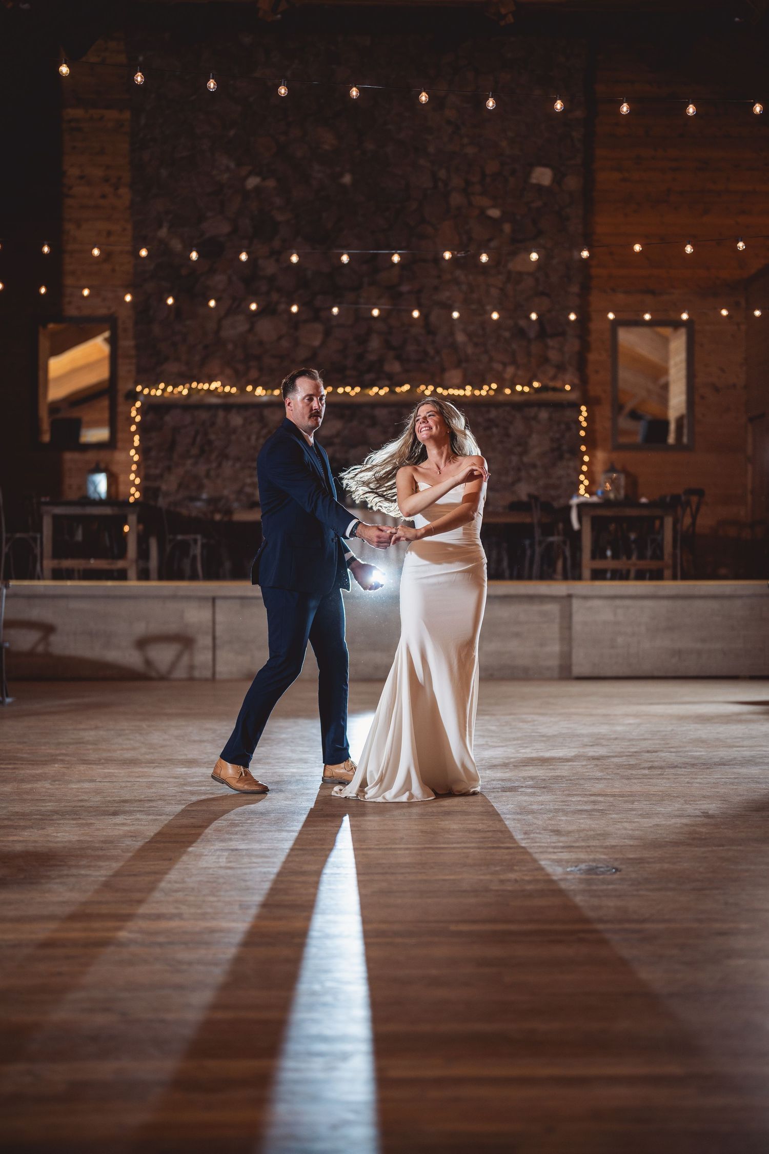 Wedding couple dances under romantic string lights in rustic indoor venue.