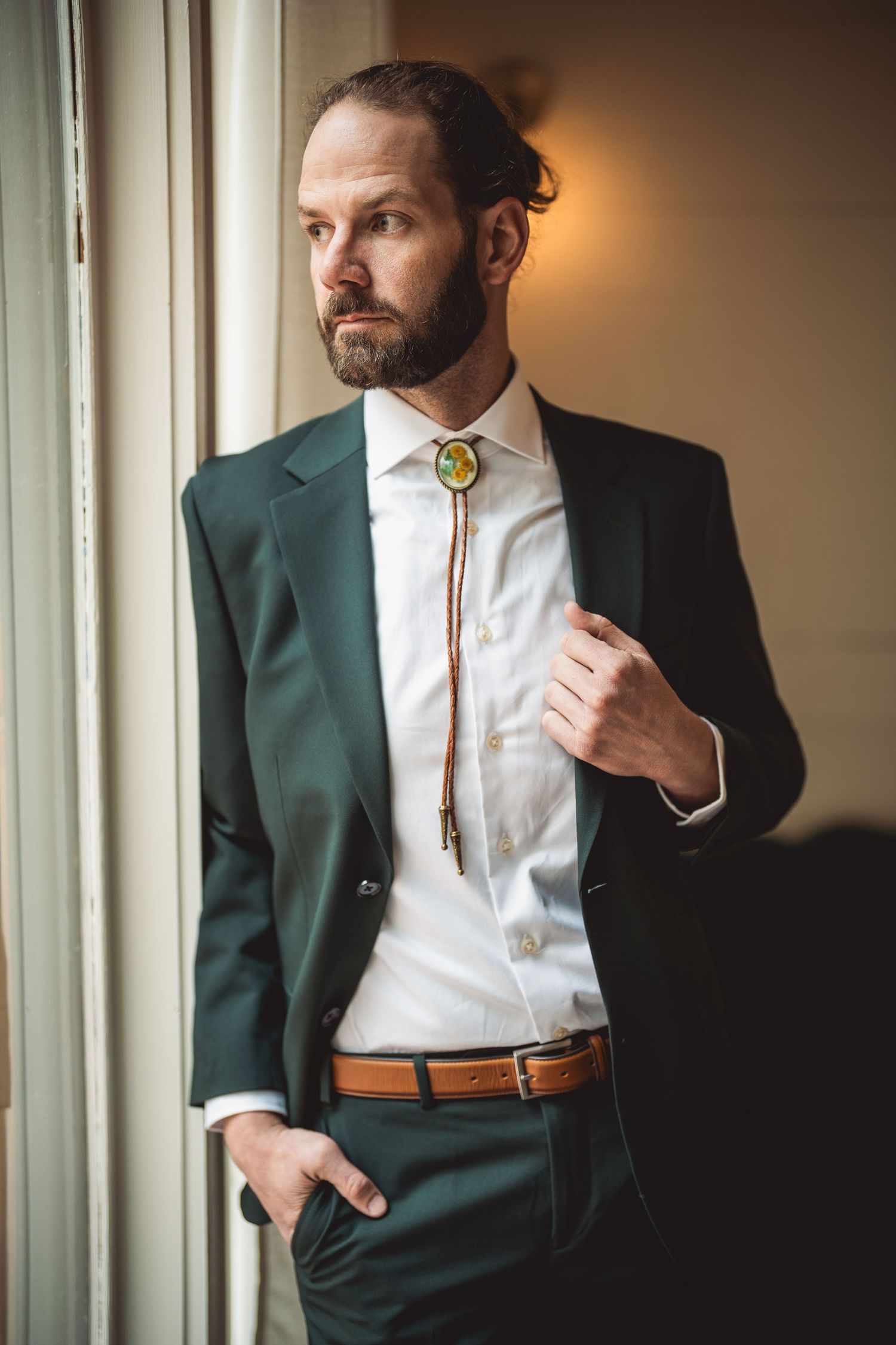 Groom in a sophisticated dark green suit posing near a window.